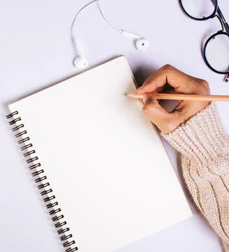 a hand writing on a note pad with earphones and glasses around it 